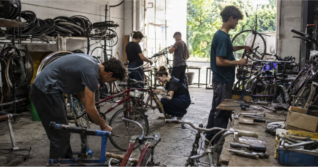 Taller mecánico con varias bicicletas usadas en reparación, jóvenes trabajando con herramientas